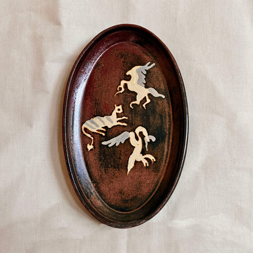 Oval oxblood and black glazed platter with three cream and grey Pompeii-inspired winged creatures